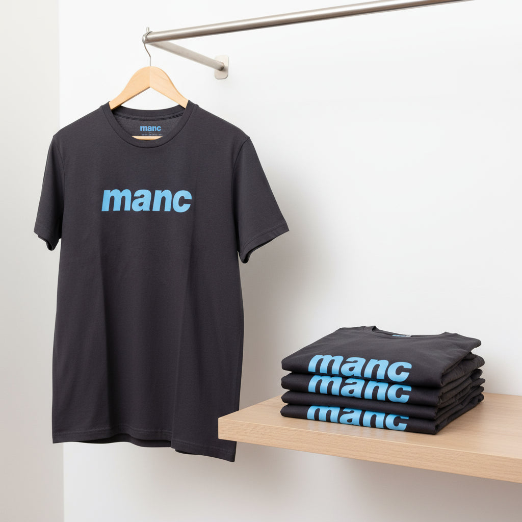 Black t-shirt with 'manc' print on a hanger and stacked on a table against a white wall.