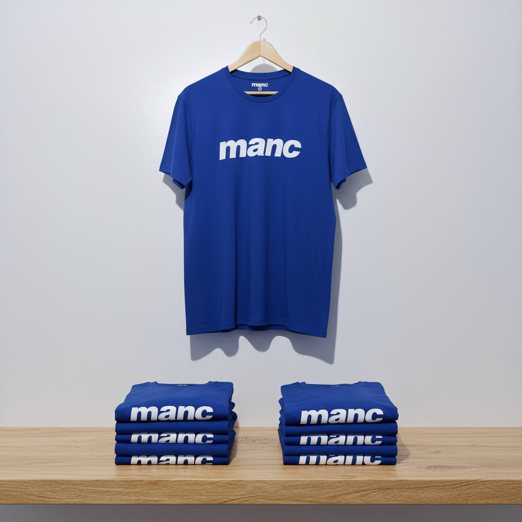 Blue t-shirt with 'manc' text on a hanger and stacked below against a white wall.