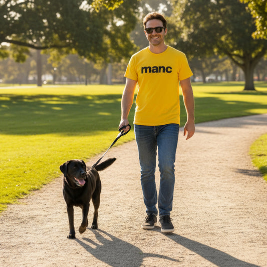 Man walking a black dog on a leash in a park wearing a yellow 'manc' t-shirt.