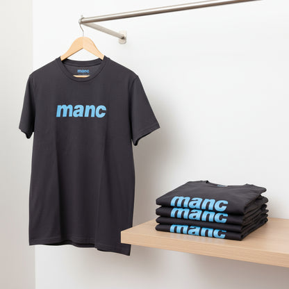 Black t-shirt with 'manc' print on a hanger and stacked on a table against a white wall.